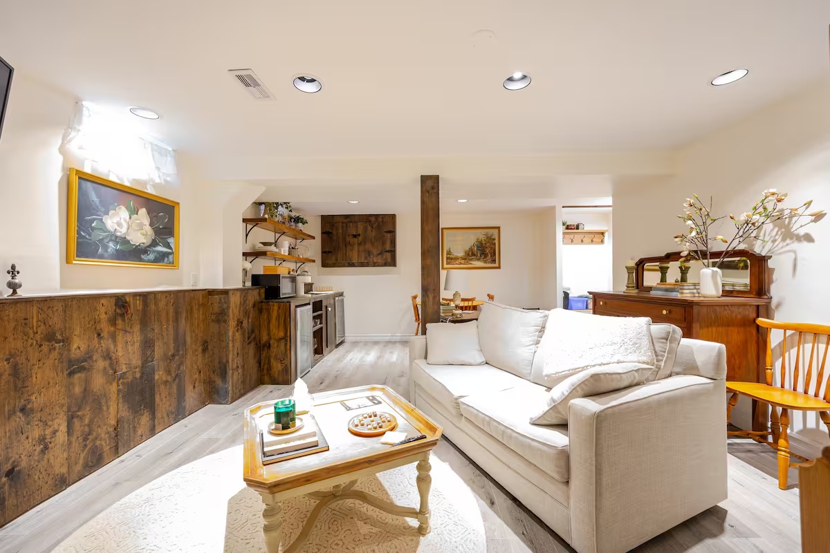 Bright lower-level living room with sofa, coffee table, kitchenette, and warm wood details